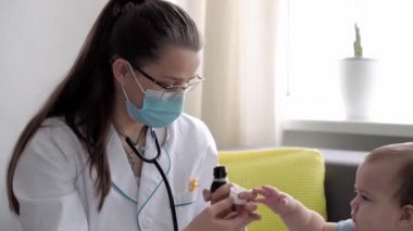 Tıp ve sağlık, pediatri, covid-19 konsepti - Kafkas Slav kökenli genç kadın hemşire ya da doktor çocuk doktorunu 8-12 aylık bebeği sarı gri kanepede muayene ediyor.
