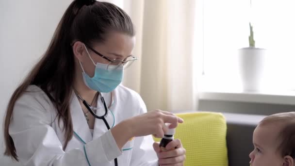 医学与健康、儿科、 covid-19概念- -近视的年轻女护士或医生、高加索斯拉夫裔儿科医生在对面的黄灰色沙发上检查8-12个月大的婴儿 — 图库视频影像