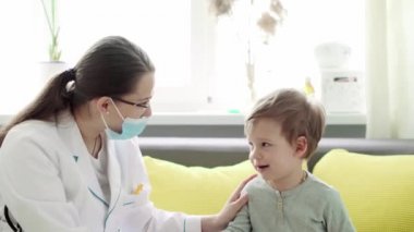 Kafkas Slav kökenli genç kadın hemşire ya da doktor çocuk doktorunun orta dereceli planı, sarı gri kanepede 2-3 yaşındaki anaokulu öğrencisi çocuğu muayene ediyor. ilaç ve sağlık, pediatri, covid-19 konsepti