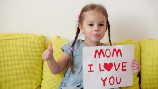在3-4岁的可爱的幼儿园小女孩手里拿着写着爱妈妈宣言的白纸。小女孩在恋爱中微笑着，手举着心形符号。浪漫的母亲节概念. — 图库视频影像