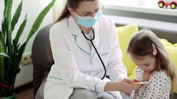 Средний план молодой кавказской славянки-медсестры или врача-педиатра измеряет температуру 3-4 года несовершеннолетней дошкольницы на желтом сером диване. медицина и здоровье, педиатрия, концепция covid-19 — стоковое видео