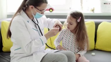 Genç kadın hemşire ya da doktor Kafkas Slav kökenli orta yaş planı sarı gri kanepede 3-4 yaş arası anaokulu öğrencisi kız çocuğuna iksir veriyor. ilaç ve sağlık, pediatri, covid-19 konsepti