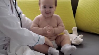 Tıp ve sağlık, pediatri, covid-19 konsepti - Kafkas Slav kökenli genç kadın hemşire ya da doktor çocuk doktorunu 8-12 aylık bebeği sarı gri kanepede muayene ediyor.