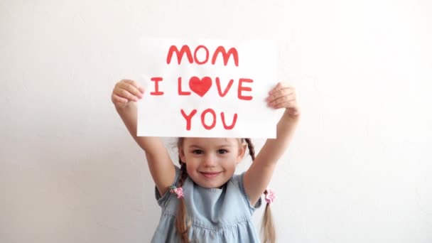 Close up cute little preschool caucasian girl 3-4 years holding white sheet with declaration of love mom. kid daughter smiling in love showing heart symbol with hands. Romantic, Mothers Day concept. — Stock Video