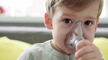 3-4 yaşlarında, sarı gri kanepenin üzerinde astım spreyi maskesi takan küçük bir anaokulu çocuğu. Bebek nebulizörle solunum terapisi görüyor. ilaç ve sağlık, pediatri, covid-19 konsepti