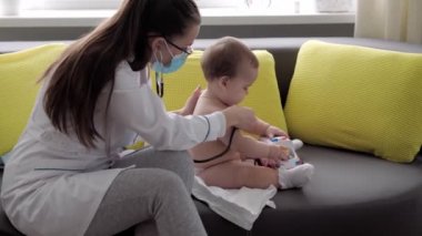 Tıp ve sağlık, pediatri, covid-19 konsepti - Kafkas Slav kökenli genç kadın hemşire ya da doktor çocuk doktorunu 8-12 aylık bebeği sarı gri kanepede muayene ediyor.