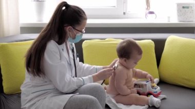 Tıp ve sağlık, pediatri, covid-19 konsepti - Kafkas Slav kökenli genç kadın hemşire ya da doktor çocuk doktorunu 8-12 aylık bebeği sarı gri kanepede muayene ediyor.