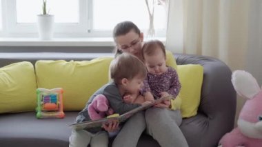 Otantik, şirin, neo-beyaz gözlüklü anne ve iki çocuk, sarı kanepe üzerinde kitap okuyorlar. Bebekleri okşayan genç bir kadın. annelik, ebeveynlik, çocukluk, hayat konsepti