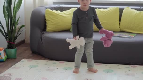 Medium plan autentiske lille førskolebarn dreng 2-3 år i sweater og jeans kaster bamser i kid room på baggrund af vindue og gul-grå sofa. barndom, liv, ægthed koncept – Stock-video