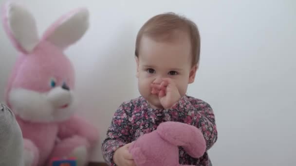 Autentisk närbild liten spädbarn flicka 1 år gammal i färgglad klänning sitter och leker med rosa nalle i barnrum på vit bakgrund inomhus. föräldraskap, barndom, livskoncept — Stockvideo