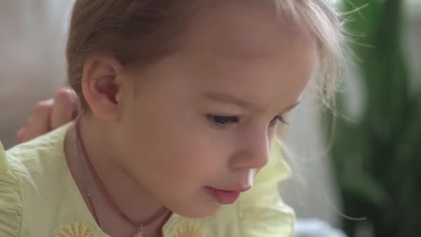 Primer plano auténtico lindo joven mujer caucásica mamá el cuidado de las trenzas para niños hija coletas en la cabeza de pelo rubio en el sofá gris amarillo. Proceso de trenzado de tejidos. Moda y belleza, concepto de maternidad — Vídeos de Stock