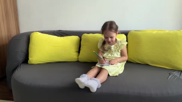 穿着黄色衣服、面带微笑的小女孩坐在家里的沙发上，用铅笔在笔记本上写字。可爱的小女孩，漂亮的脸蛋在灰色的沙发上。童年、远距离学习概念 — 图库视频影像