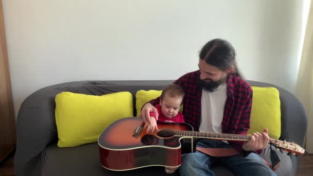 Mann mit schwarzem Bart lange Haare in Rock-Stil verbringen Zeit Kleinkind Gitarre spielen zu Hause. Der Vater bringt seiner kleinen Tochter das Spielen von Musikinstrumenten bei und genießt den Musikunterricht im Haushalt. Familienkonzept — Stockvideo