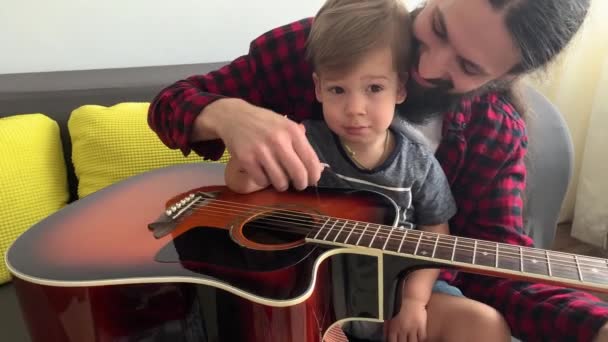 ロックスタイルの幸せな男は居心地の良い家でギターを弾く幼児と時間を過ごす。父は小さな息子に家庭内音楽のレッスンを楽しむ楽器を教える。家族教育人生 — ストック動画