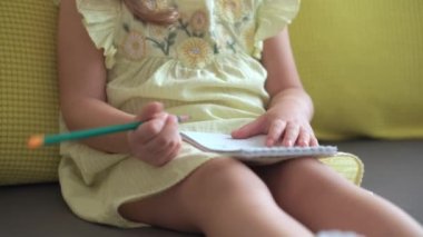 Sarı elbiseli, gülümseyen çocuk evdeki kanepede oturan kalemle yazıyor. Gri kanepede güzel yüzlü tatlı bir bayan çocuk. çocukluk, uzaklık öğrenme kavramı