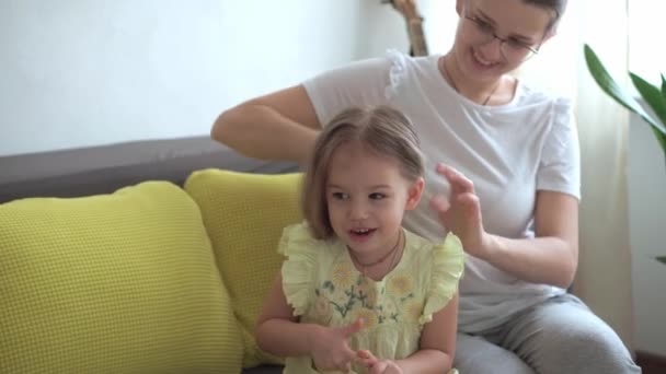 Chiudere autentico carino giovane donna caucasica mamma prendersi cura di trecce bambino figlia trecce sulla testa dei capelli biondi su divano grigio giallo. Processo di intreccio trecce. Moda e bellezza, concetto di maternità — Video Stock