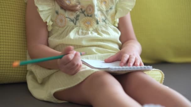 Porträtt Rolig liten flicka i gul klänning leende barn skriver med penna i anteckningsbok sitter på soffan hemma. söt unge kvinna med vackert ansikte på grå soffa. barndom, distansundervisning — Stockvideo