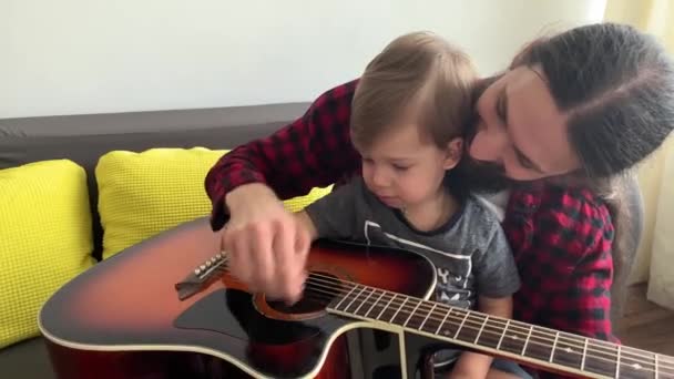 Hombre feliz en estilo rock pasar tiempo con un niño pequeño tocando la guitarra en casa acogedora. padre enseñar a su hijo pequeño a tocar el instrumento musical disfrutando de la lección de música doméstica. familia, educación, vida — Vídeo de stock