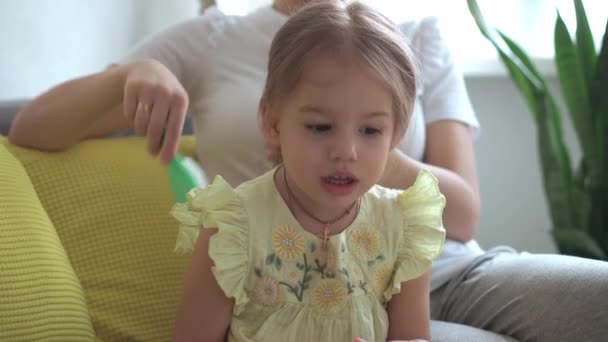 Primer plano auténtico lindo joven mujer caucásica mamá el cuidado de las trenzas para niños hija coletas en la cabeza de pelo rubio en el sofá gris amarillo. Proceso de trenzado de tejidos. Moda y belleza, concepto de maternidad — Vídeo de stock