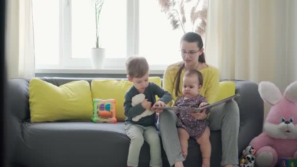 Authentic close up cute neo caucasian spectacled mom with 2 children boy and girl on gray yellow sofa reading book. Young woman caressing infant baby. motherhood, parenthood, childhood, life concept — Stock Video