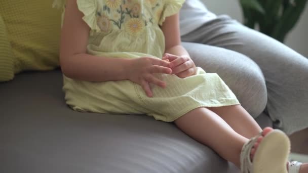 Primer plano auténtico lindo joven mujer caucásica mamá el cuidado de las trenzas para niños hija coletas en la cabeza de pelo rubio en el sofá gris amarillo. Proceso de trenzado de tejidos. Moda y belleza, concepto de maternidad — Vídeos de Stock