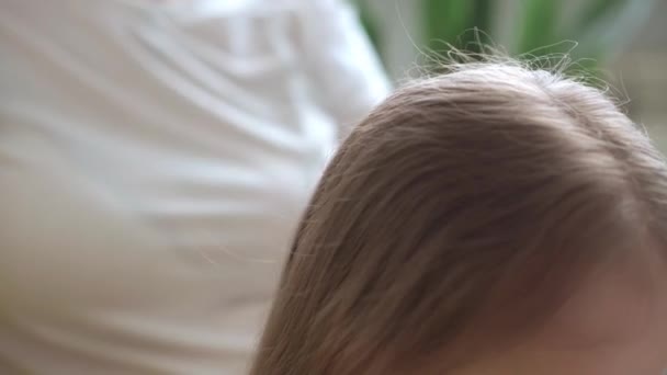Close up autêntico bonito jovem caucasiano mãe cuidar de crianças tranças filha tranças no cabelo loiro cabeça no sofá cinza amarelo. Processo de trançar tecelagens. Moda e beleza, conceito de maternidade — Vídeo de Stock