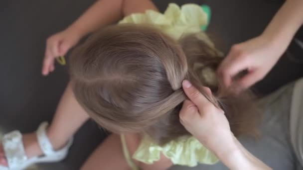 Primer plano auténtico lindo joven mujer caucásica mamá el cuidado de las trenzas para niños hija coletas en la cabeza de pelo rubio en el sofá gris amarillo. Proceso de trenzado de tejidos. Moda y belleza, concepto de maternidad — Vídeos de Stock