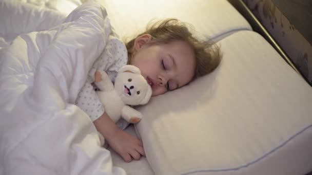 真真切切地关闭可爱的高加索幼儿3-4岁小女孩与泰迪熊睡在舒适的白色床上。幼儿保育、熟睡婴儿、童年、父母身份、人生观 — 图库视频影像