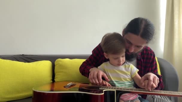 Glücklicher Mann im Rockstil verbringt Zeit mit Kleinkind, das zu Hause Gitarre spielt. Vater bringt kleinem Sohn das Spielen von Musikinstrumenten bei und genießt den häuslichen Musikunterricht. Familie, Bildung, Leben — Stockvideo