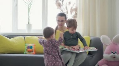 Otantik, şirin, neo-beyaz gözlüklü anne ve iki çocuk, sarı kanepe üzerinde kitap okuyorlar. Bebekleri okşayan genç bir kadın. annelik, ebeveynlik, çocukluk, hayat konsepti