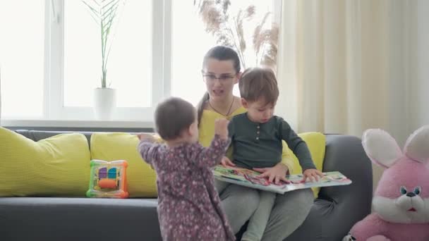 Autentyczne zbliżenie cute neo kaukaski spektakularne mama z 2 dzieci chłopiec i dziewczyna na szary żółty sofa czytania książki. Młoda kobieta pieści niemowlę. macierzyństwo, rodzicielstwo, dzieciństwo, koncepcja życia — Wideo stockowe