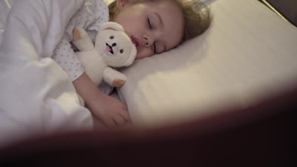 3-4 yıl boyunca tatlı beyaz bir yatakta oyuncak ayıyla tatlı tatlı tatlı uyuyan, beyaz, küçük beyaz bir anaokulu çocuğunun otantik yakın çekimi. Çocuk bakımı, Uyuyan Bebek, Çocukluk, Ebeveynlik, Yaşam Konsepti — Stok video