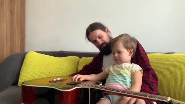 Hombre feliz en estilo rock pasar tiempo con un niño pequeño tocando la guitarra en casa acogedora. padre enseñar a su hijo pequeño a tocar el instrumento musical disfrutando de la lección de música doméstica. familia, educación, vida — Vídeos de Stock