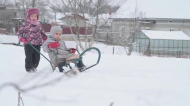 Tatil, oyun, aile kavramı - yavaş çekim otantik iki mutlu anaokulu çocuğu kardeşler şapkalar ve eldivenler takıp birbirlerini yuvarlıyorlar. Soğuk mevsimde kar yağışı, kış parkında açık havada.