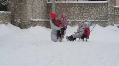 Tatil, oyun, aile kavramı, yavaş çekim otantik üç mutlu anaokulu çocuğu, genç Wonan annesi kızakla kayan ve karla oynayan kardeşleri. Soğuk mevsimde kar yağışı açık havada kış bahçesinde