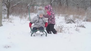 Tatil, oyun, aile kavramı - yavaş çekim otantik iki mutlu anaokulu çocuğu kardeşler şapkalar ve eldivenler takıp birbirlerini yuvarlıyorlar. Soğuk mevsimde kar yağışı, kış parkında açık havada.