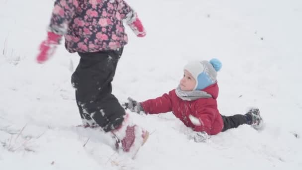 Wakacje, gra, koncepcja rodziny - slo-mo autentyczne Dwa szczęśliwe przedszkolaki rodzeństwo dzieci zjeżdżają ze wzgórza i pomagają sobie nawzajem wspiąć. opady śniegu w zimnym sezonie pogoda w parku zimowym na zewnątrz — Wideo stockowe