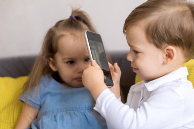 Mutlu ve üzgün küçük anaokulu kardeşleri küçük erkek ve kız kardeş sarı gri kanepede video konferansı hakkında konuşuyorlar. Oğlan, üzgün kızın resimlerini akıllı telefonla çekiyor. Çocukluk, teknoloji konsepti