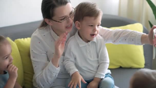 Kaukasiska familj små barn flicka pojke och mamma talar i videokonferens via smartphone tillsammans i vardagsrummet hemma med glädje leende. aktivitetsteknik livsstil mobiltelefon användning koncept — Stockvideo