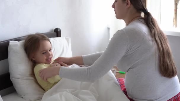 La enfermera o el médico jóvenes caucásicos examina a la niña mide la temperatura con el termómetro debajo del brazo en la cama blanca. Mamá cuida a un niño enfermo. medicina y salud, maternidad, concepto covid-19 — Vídeo de stock