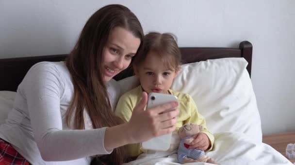Kaukasiske familie små børn pige og ung kvinde mor taler i videokonference ved smartphone sammen derhjemme med lykke smil på hvid seng. teknologi, livsstil, mobiltelefonbrugskoncept – Stock-video