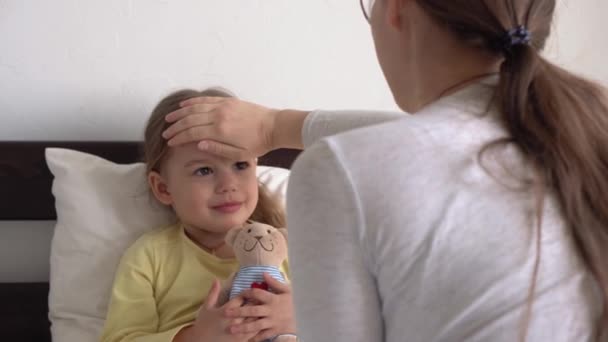 Młoda kaukaska kobieta pielęgniarka lub lekarz bada dziewczynkę mierzy temperaturę dłonią czoła na białym łóżku. Mama zajmuje się chorym dzieckiem. medycyna i zdrowie, macierzyństwo, koncepcja covid-19 — Wideo stockowe