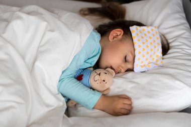 Otantik portre, beyaz yatakta alnına baskı uygulayan mavi elbiseli, beyaz, beyaz, şirin, beyaz bir anaokulu bebeği. Çocuk öğle yemeği vakti dinleniyor. bakım, ilaç ve sağlık, çocukluk, yaşam konsepti