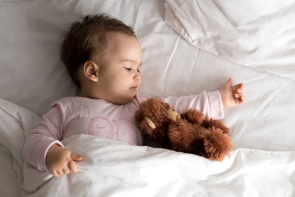 Retrato autêntico bonito caucasiano pequeno bebê gordinho menina ou menino em rosa sonolento ao acordar com ursinho olhando para a câmera na cama branca. Cuidados infantis, Infância, Paternidade, conceito de estilo de vida — Fotografia de Stock