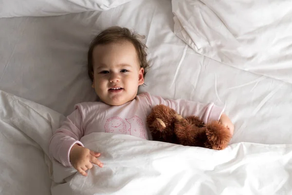 Hiteles portré aranyos kaukázusi kis csecsemő pufók kislány vagy fiú rózsaszín álmos ébredés után maci nézi kamera fehér ágyban. Gyermekgondozás, Gyermekkor, Szülőkor, életmód koncepció — Stock Fotó