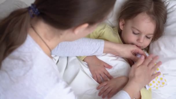 Close-up jonge blanke vrouw verpleegkundige of arts kinderarts onderzoekt baby meisje zet kompres op het voorhoofd op wit bed. Mama zorgt voor ziek kind. geneeskunde en gezondheid, moederschap, covid-19-concept — Stockvideo