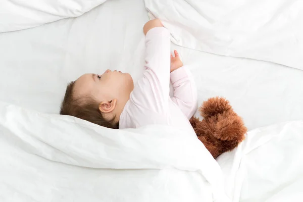 Authentique portrait mignon caucasien petit bébé joufflu bébé fille ou garçon en somnolence rose au réveil avec ours en peluche regardant la caméra dans un lit blanc. garde d'enfants, enfance, parentalité, concept de style de vie — Photo