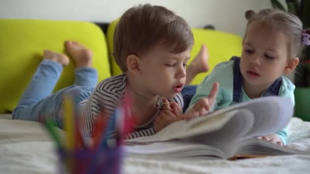 Przyjaźń, rodzina, edukacja - Dwoje uśmiechniętych przedszkolaków, maluchy czytają na żółtym łóżku dużą ciekawą książkę bajek. Rodzeństwo małe bliźniaki czytelnik zabawy, szczęśliwe dzieci na kwarantannie w domu — Wideo stockowe