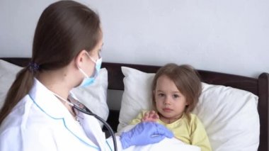 Otantik, şirin, anaokulu hasta kızı beyaz yatakta kameraya bakıyor. Kafkas genç kadın hemşire ya da doktor çocuk doktoru hastanede bebeği muayene eder. tıp ve sağlık, covid-19 konsepti