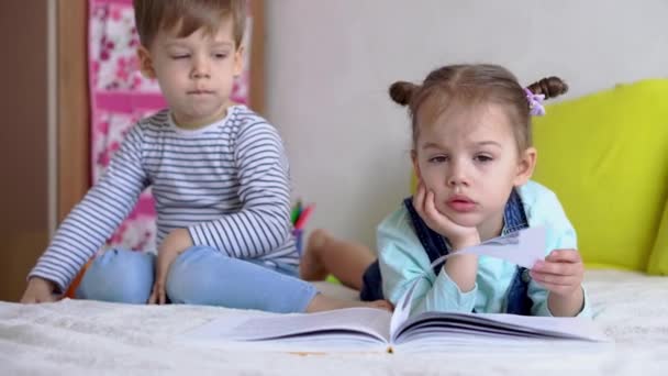 Vänskap, familj, utbildning - Två leende preshool, småbarn läser en stor intressant bok om sagor på gul säng. Syskon små tvillingar läsare ha kul, glada barn på karantän hemma — Stockvideo